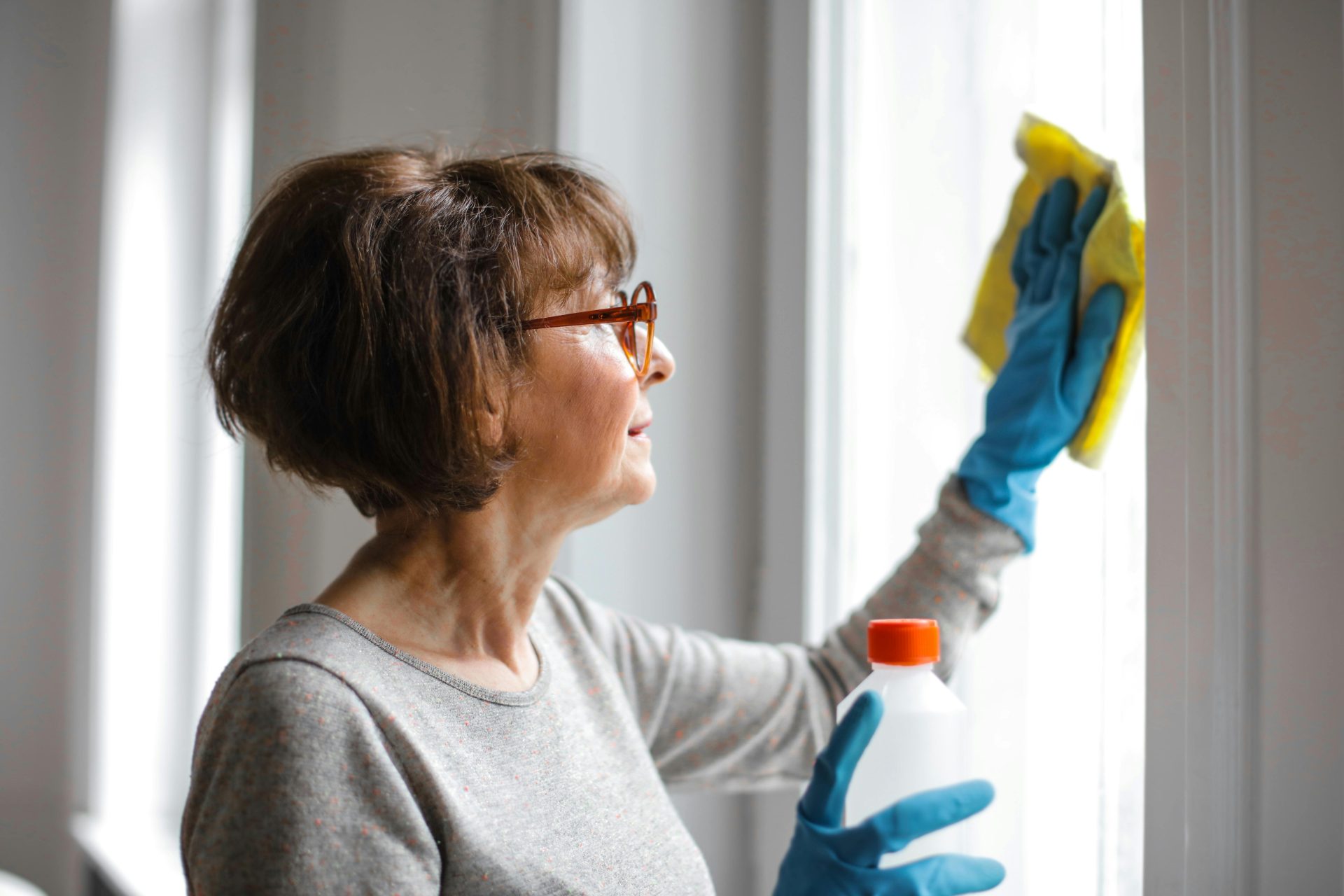 Ältere Frau reinigt Fenster im Innenbereich mit Handschuhen und Reiniger.