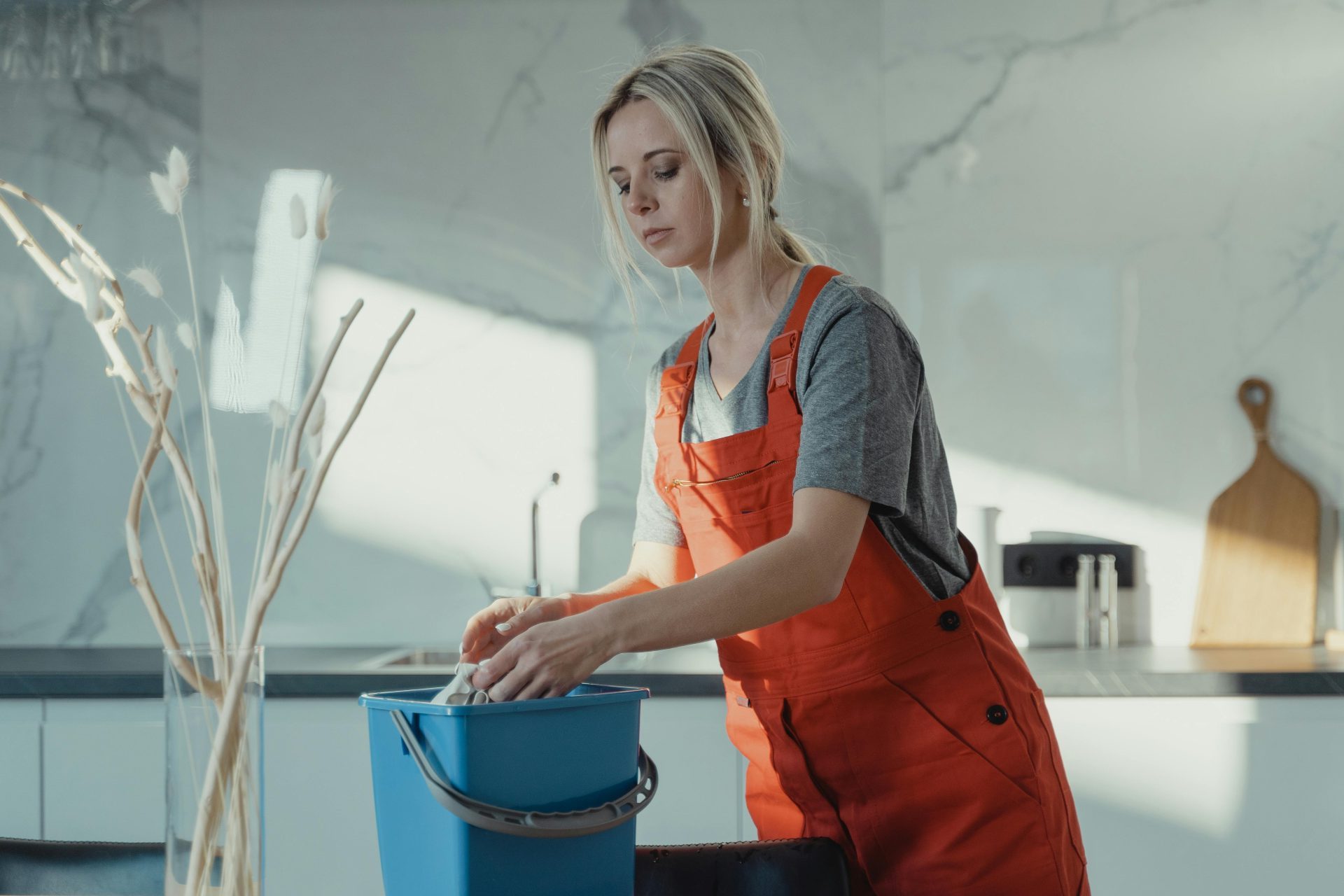 Eine Frau in grauem Hemd und rotem Overall reinigt eine Küchentheke mit einem blauen Eimer und einem weißen Lappen. Modernes Ambiente.